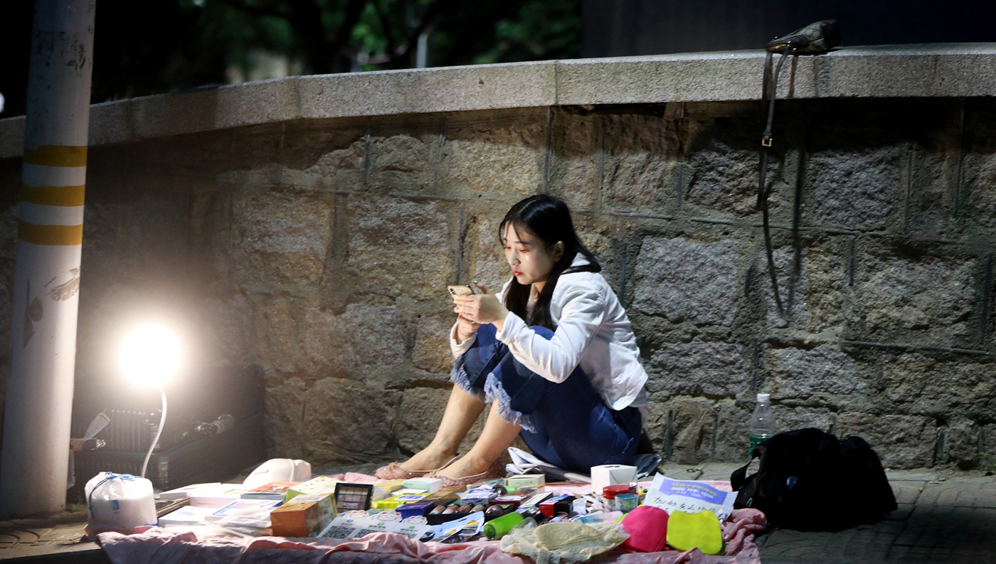 一位女大学生在摆摊的时候刷手机,不管你买不买,我和货品就在这里,不